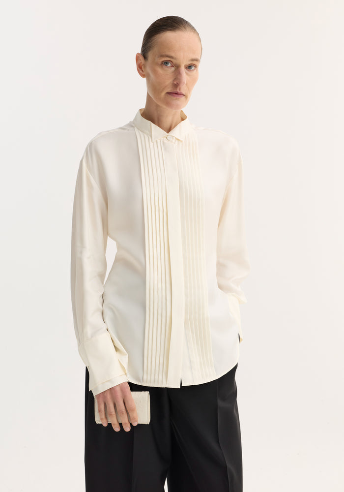 Shaped tuxedo silk shirt in cream on model, featuring a tuxedo-style pleated front, stand-up collar, double cuffs, paired with black trousers.