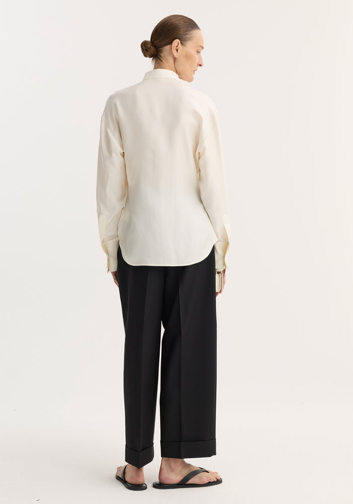 Person wearing Shaped tuxedo silk shirt with stand-up collar, double cuffs, pleated front, over loose black trousers and black sandals, hair in low bun.