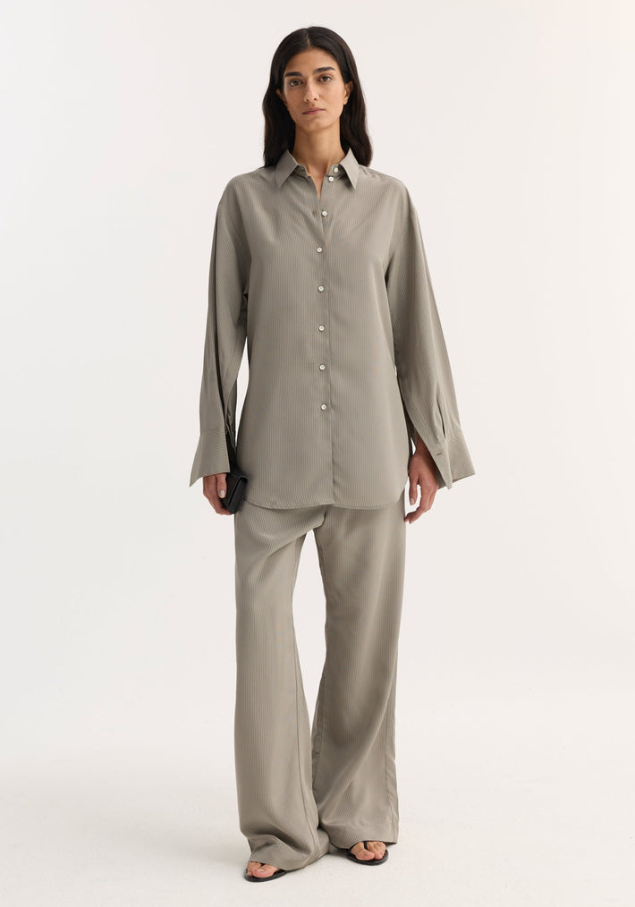 Woman wearing Relaxed shaped shirt in light grey stripe, featuring a loose fit, button-down design, oversized cuffs, and wide-leg trousers, holding a black clutch.