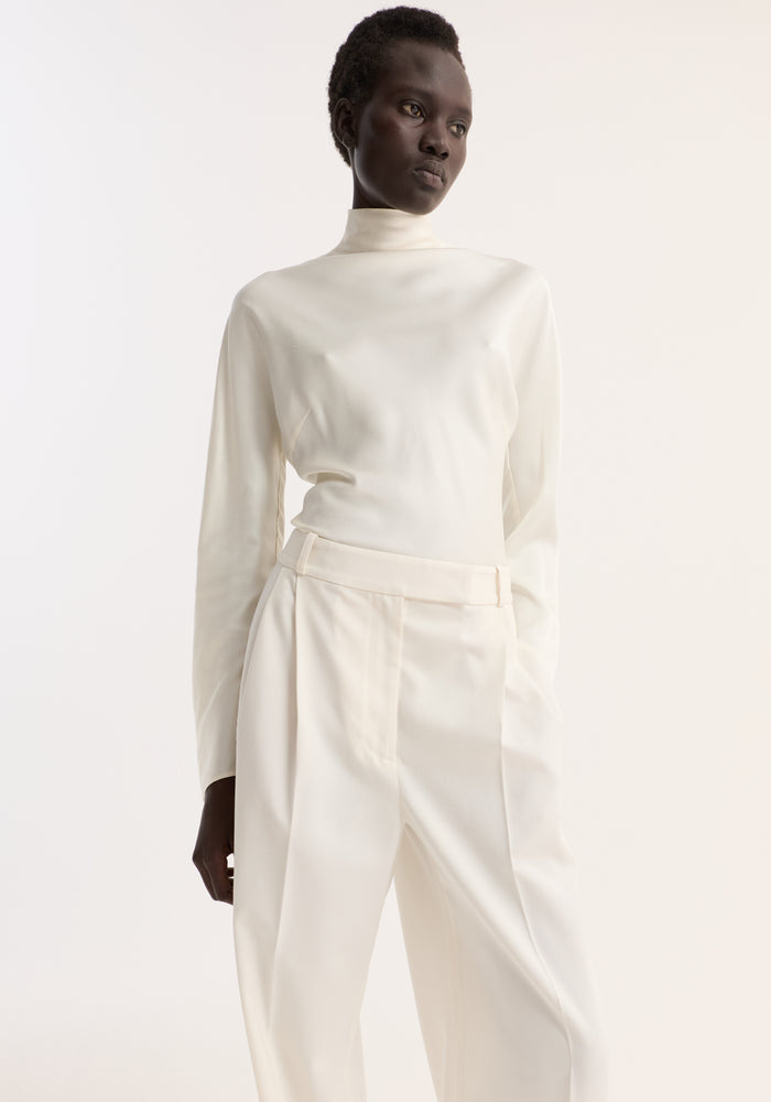 Woman wearing a fitted Satin waisted top with long sleeves, paired with high-waisted pleated trousers, posing with hand in pocket, exuding minimalist elegance.