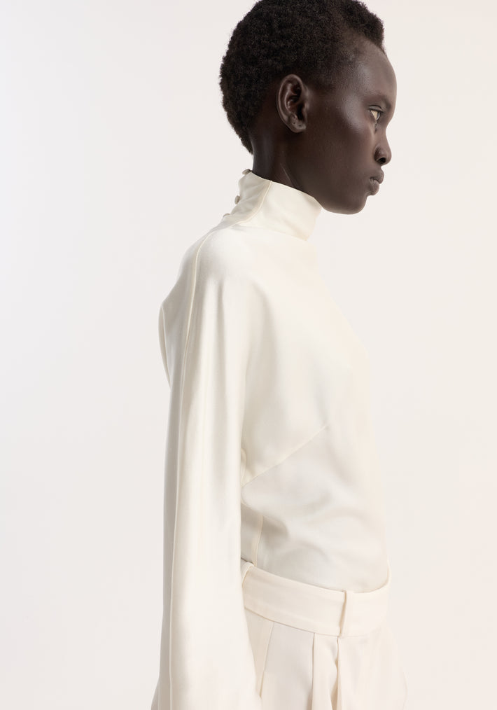 Woman in a minimalist all-white outfit showcasing the Satin waisted top in cream, featuring a high-neck and button-fastened collar, paired with pleated trousers.