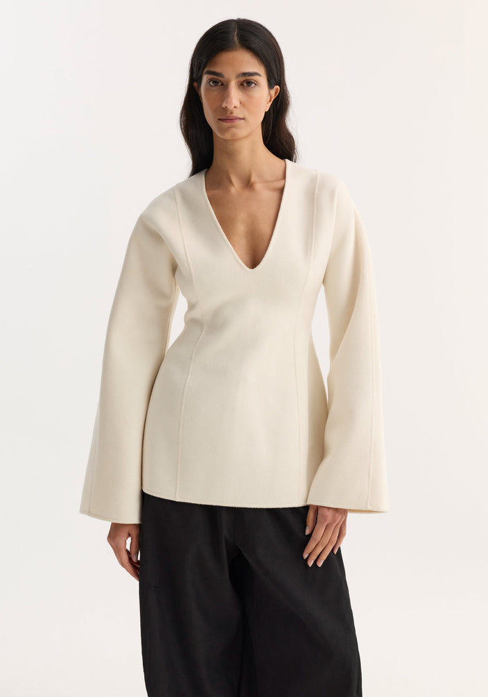 Woman wearing Sculptural long sleeve top | off white with voluminous bell sleeves and V-neck, paired with loose black trousers; architectural silhouette emphasizes the defined waist.