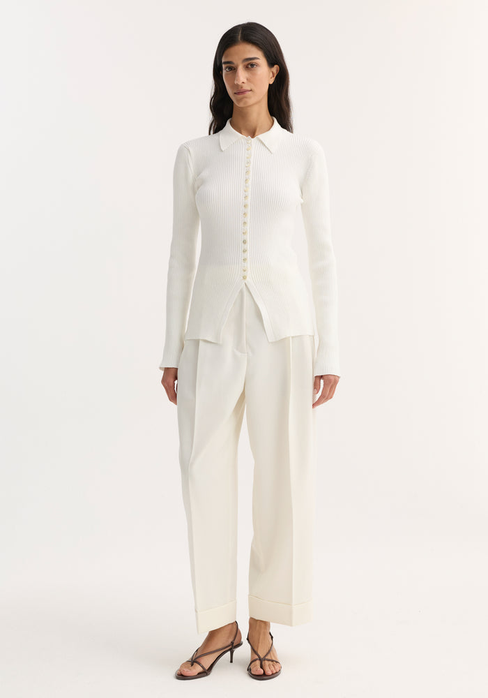 Model wears Elongated Rib Cardigan in off white, featuring a collar, ribbed texture, and front buttons ending at a hem slit, paired with high-waisted trousers.