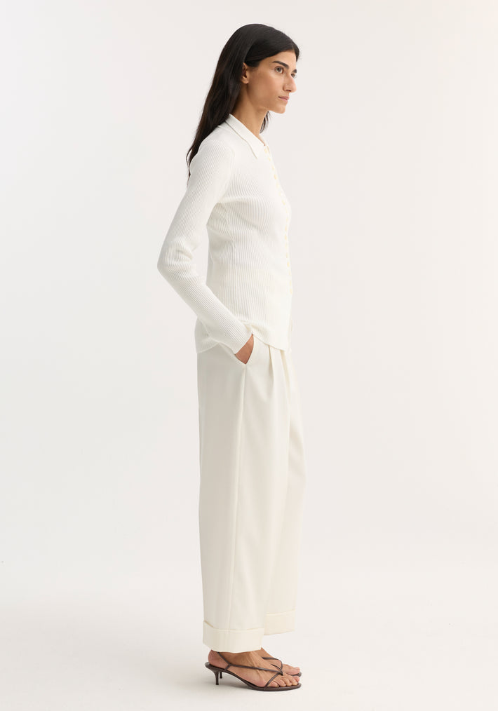 Model in profile wearing an Elongated rib cardigan in off white, showcasing button closures, collar, and ribbed knit texture, paired with wide-leg trousers and sandals.