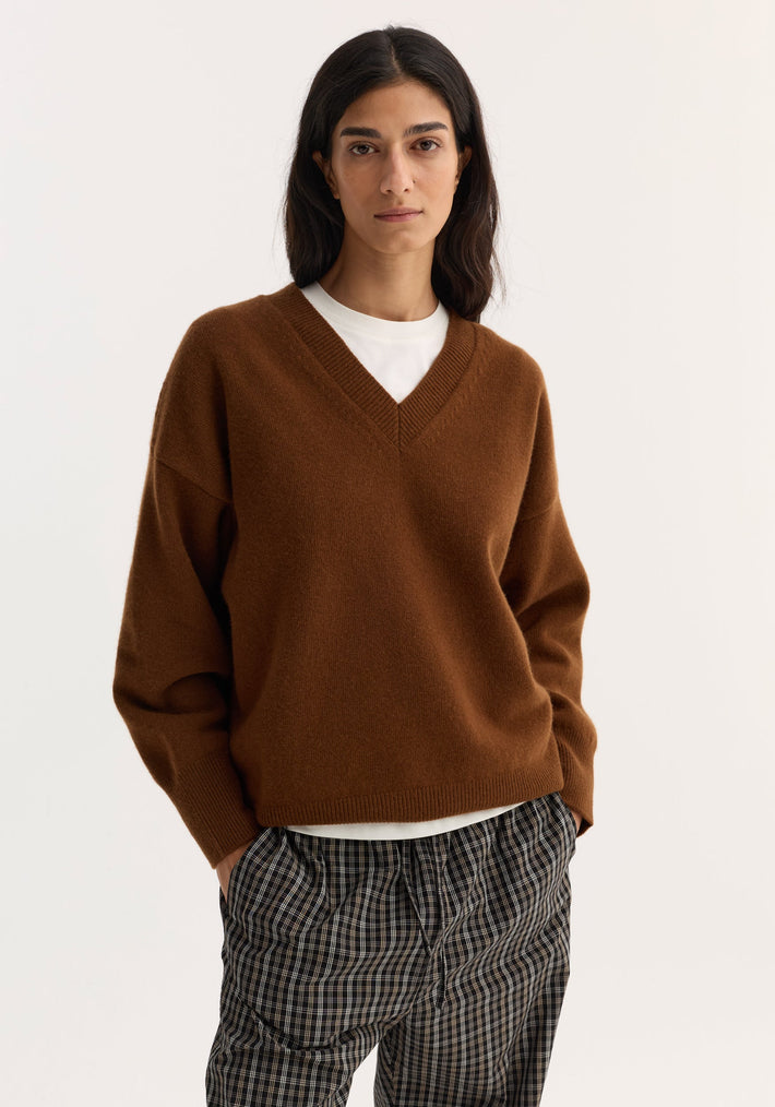 Woman wearing V-neck knitted jumper in rust, featuring a relaxed fit and dropped shoulders, paired with black-and-white checked trousers. Hands in pockets, neutral expression.