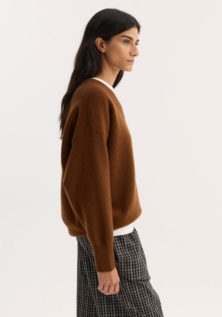 Side-profile of a person wearing a V-neck knitted jumper in rust, featuring a relaxed fit, long sleeves, and drop shoulders, paired with checkered trousers.