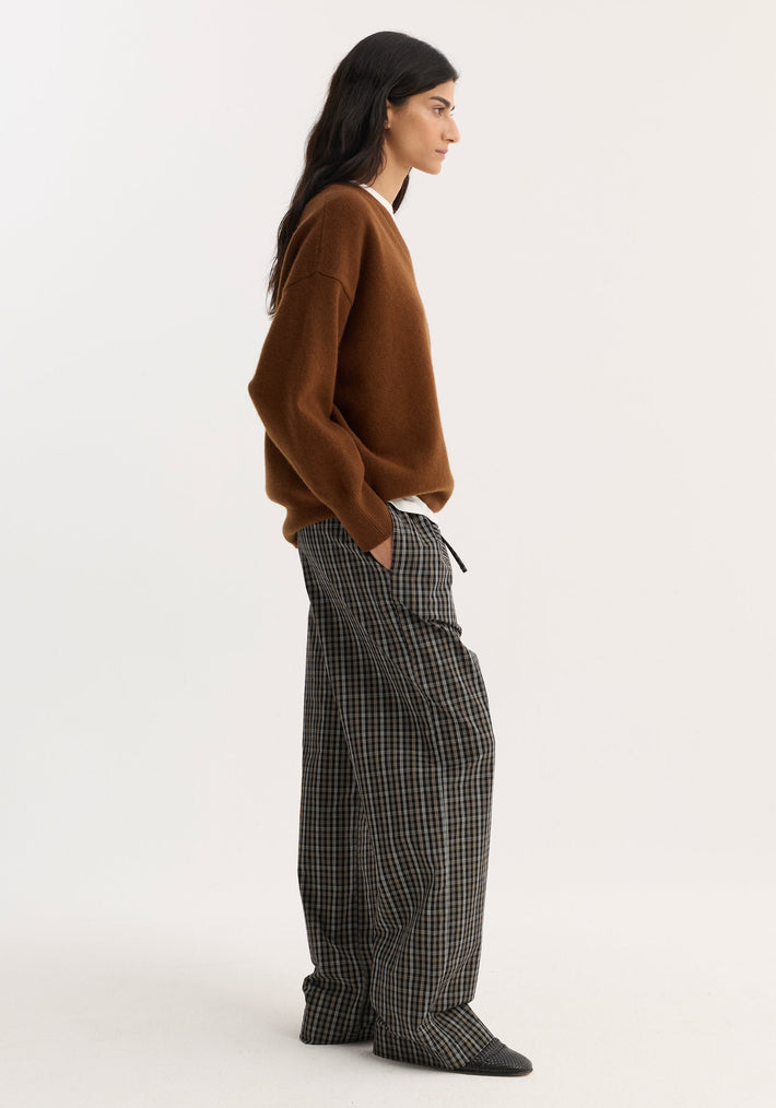 Side-profile of a person wearing a V-neck knitted jumper in rust, featuring a relaxed fit and drop shoulders, styled with checkered trousers and black flats.