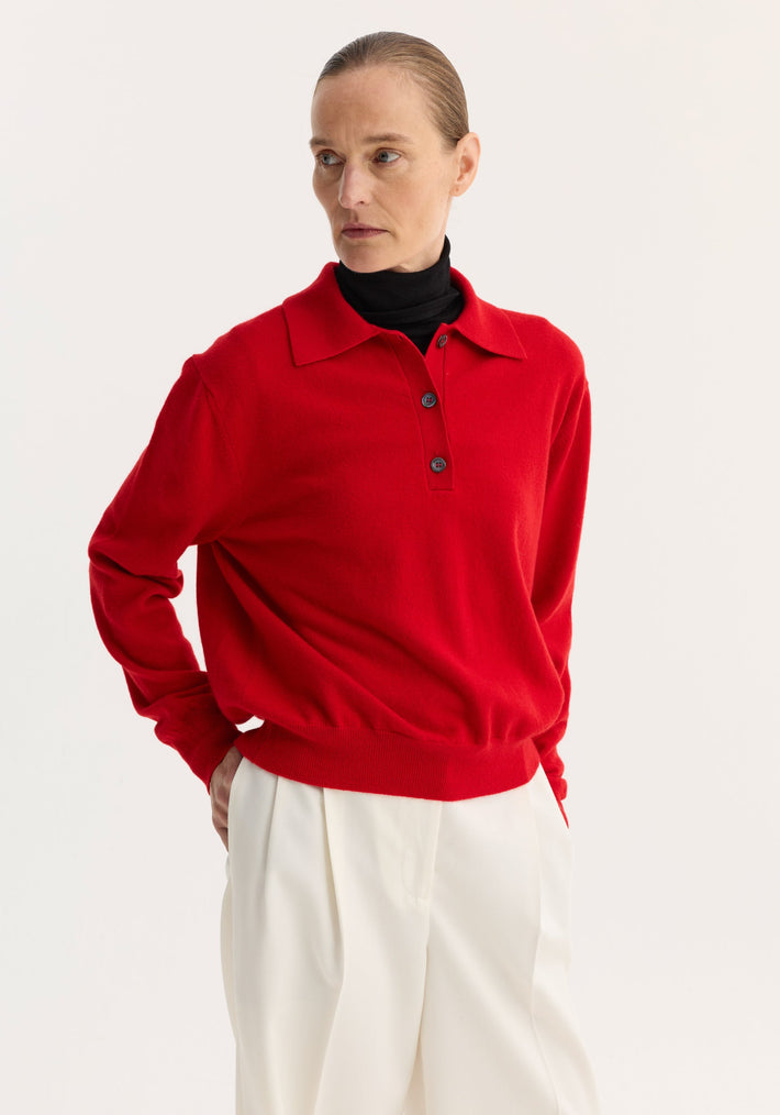 Wool cashmere polo in red, worn by a woman layered over a black turtleneck, features a structured collar and three-button front, paired with white trousers.