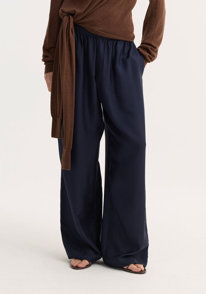 Model wearing wide-leg silk trousers | night, showing loose fit with elastic waistband and drawstring, paired with brown thong sandals.