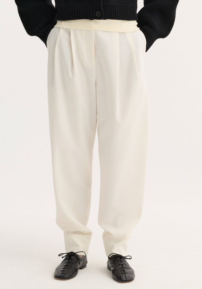 Barrel leg trousers in off-white wool blend, high-waisted with single pleat, tapering to ankle cuffs, paired with black flats against a light background.
