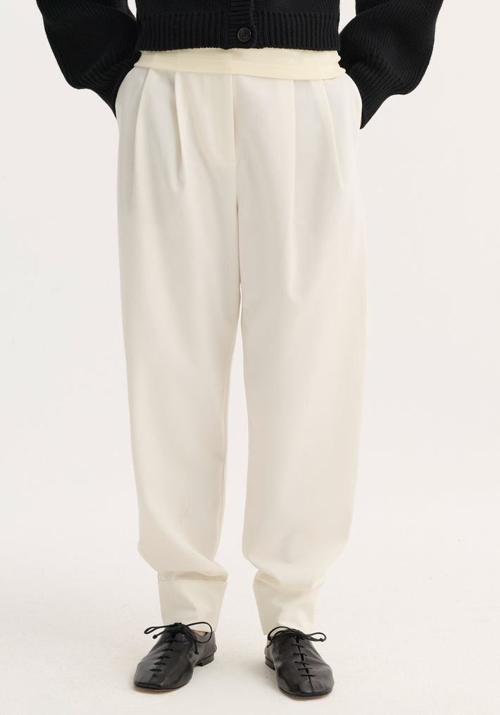 Barrel leg trousers in off-white wool blend, high-waisted with single pleat, tapering to ankle cuffs, paired with black flats against a light background.