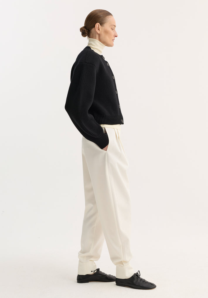 Barrel leg trousers in off-white, featuring a high-rise, double pleats, and ankle cuffs, worn by a model paired with a black cardigan and lace-up flats.