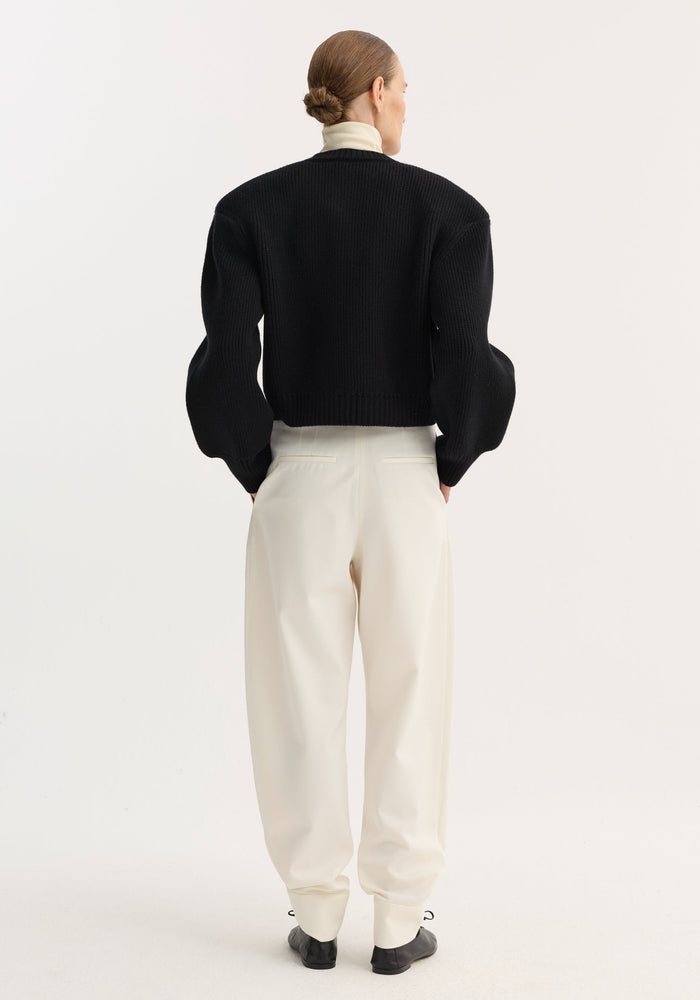 Person wearing Barrel leg trousers | off white, featuring a high-rise, wide curved legs, single pleat, and ankle cuffs, styled with a black sweater and black shoes.