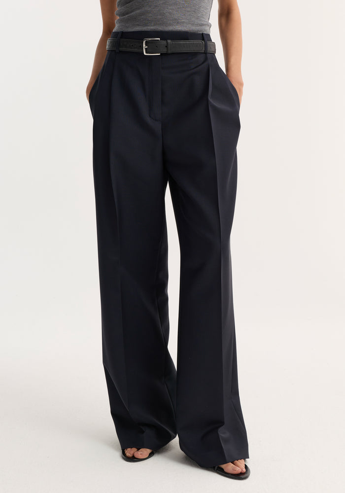 Mid-waist tailored trousers in navy, featuring a high-waisted fit, wide-leg cut, sharp pleats, and a slim black belt with a silver buckle.