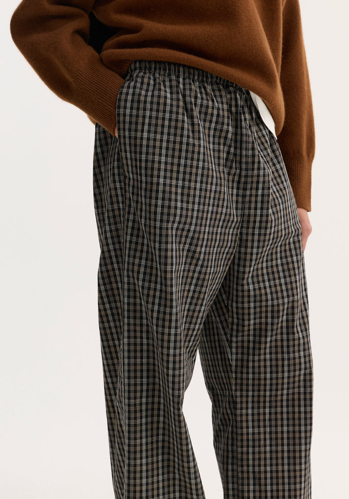 Close-up of relaxed fit trousers in black and brown check, featuring a drawstring waistband and side pockets, styled with a rust-brown knit sweater.