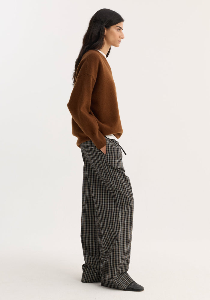 Profile view of model in relaxed fit trousers with black/brown check, featuring wide legs, drawstring waistband, and side seam pockets, styled casually with a rust sweater.