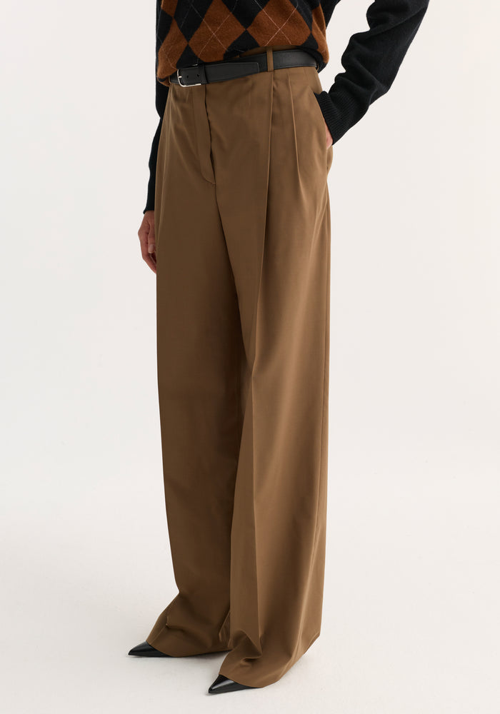 Mid-waist tailored trousers in mocha with wide-leg design, front pleats, and a black belt. Styled with black pointed-toe shoes, argyle sweater visible.