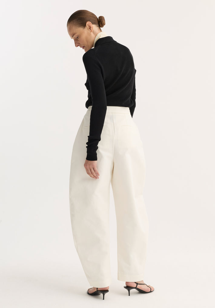 Woman wearing Sculptural trousers | paper white with high waist and balloon leg, paired with a fitted black top and open-toe sandals, hair in a bun.