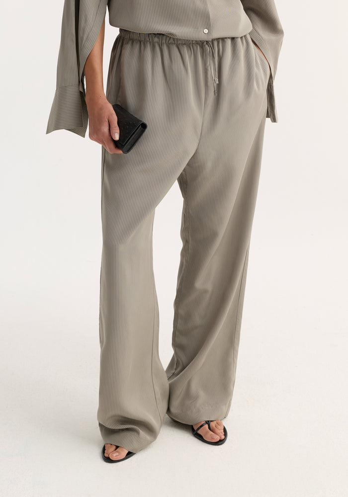 Relaxed fit trousers in light grey stripe, featuring a wide leg, drawstring waistband, and side seam pockets, styled with black sandals and a leather clutch.