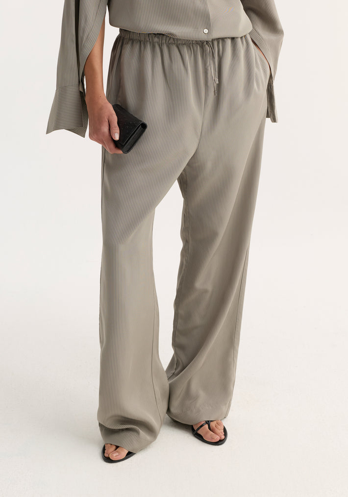 Relaxed fit trousers in light grey stripe, featuring a wide leg, drawstring waistband, and side seam pockets, styled with black sandals and a leather clutch.
