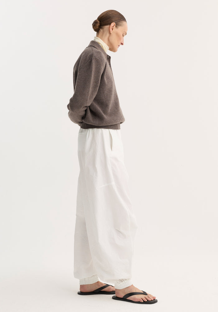 Model wearing Balloon track pants in optic white, featuring crinkled fabric, elasticated waistband, ankle cuffs, and front welt pockets, styled with a turtleneck and pullover.