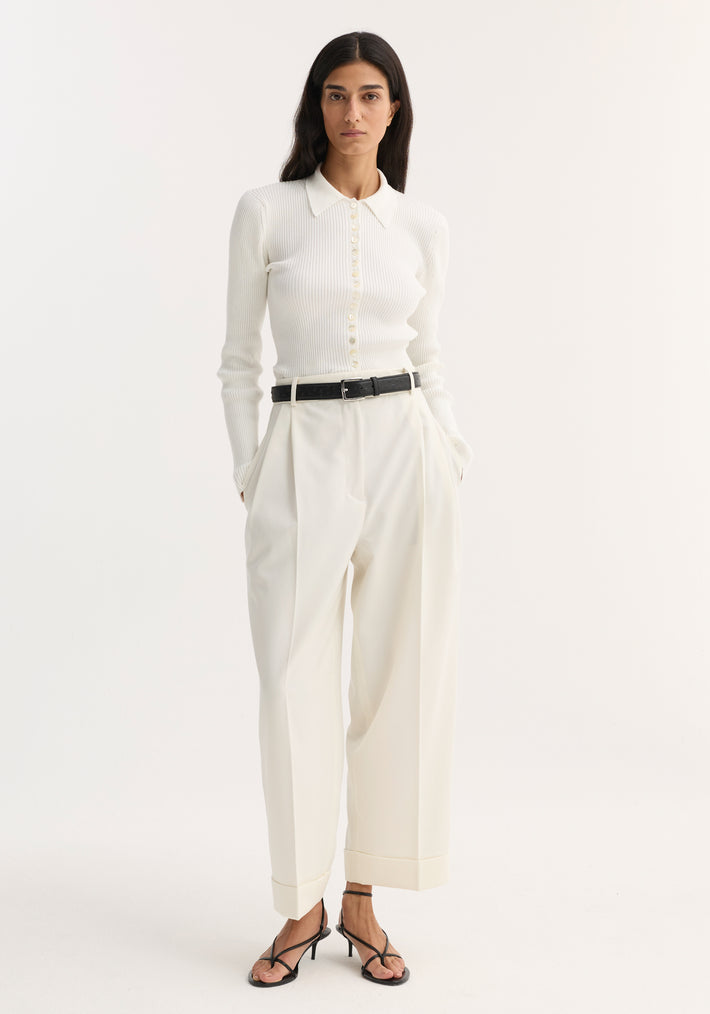Woman wearing high-waist turn-up trousers with sharp pleats, paired with a ribbed knit top, black belt, and strappy sandals.