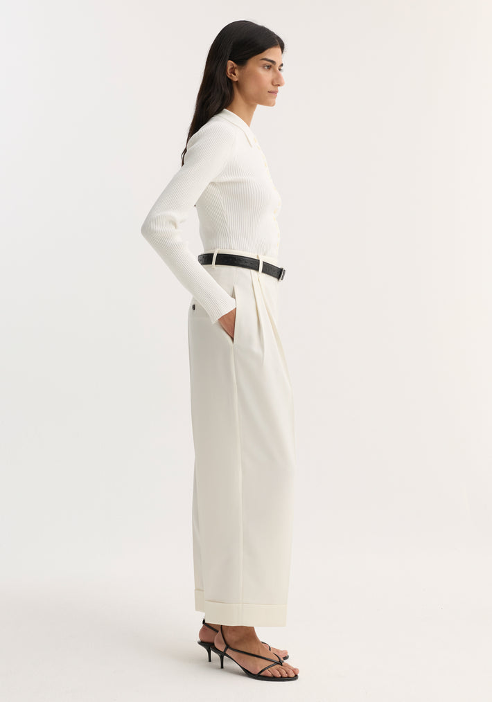 Woman in profile wearing high-waist turn-up trousers, off white, with pleats and turned-up hems, paired with a ribbed top and black sandals.