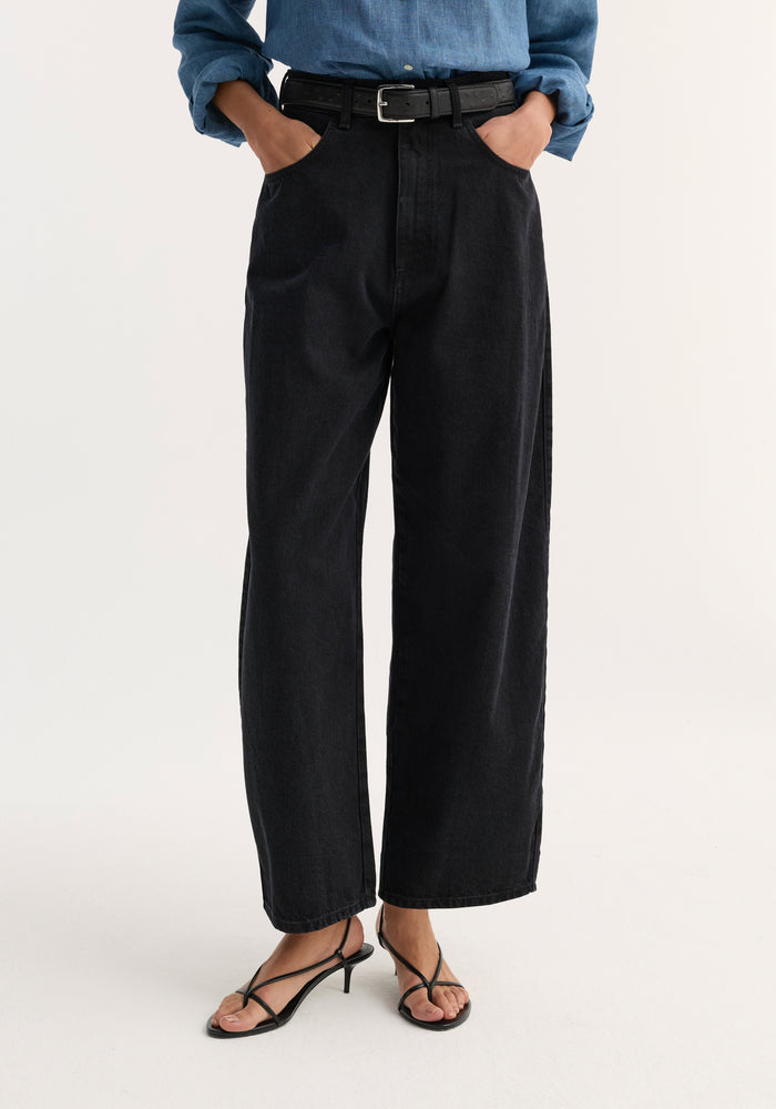Barrel leg denim in black, featuring a curved, slightly cropped barrel-leg silhouette. Worn with a leather belt, strappy heeled sandals, hands in pockets.