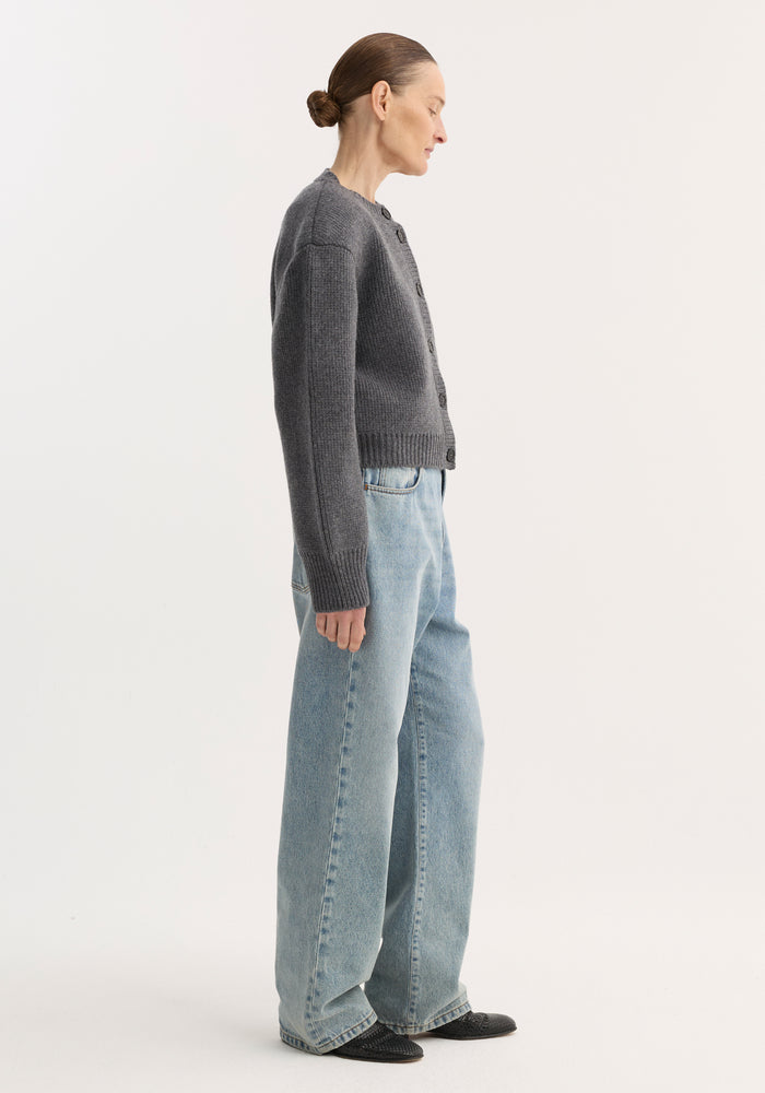 Side-profile of a person in baggy fit denim, classic blue, low-waist, wide leg, paired with a gray cardigan and black pointed-toe shoes.