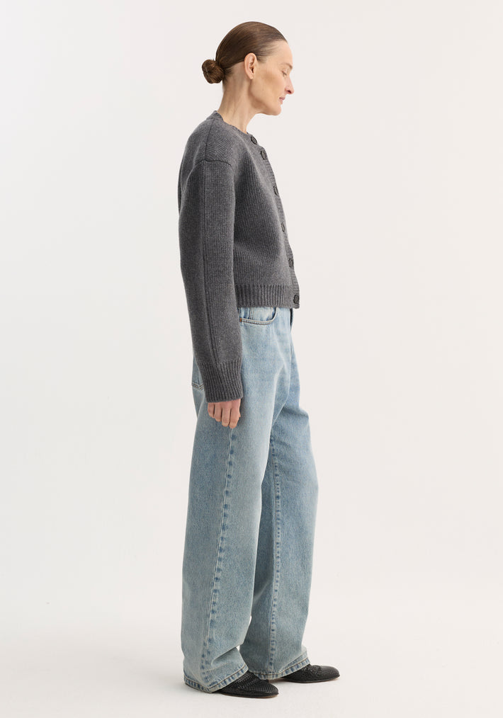 Side-profile of a person in baggy fit denim, classic blue, low-waist, wide leg, paired with a gray cardigan and black pointed-toe shoes.