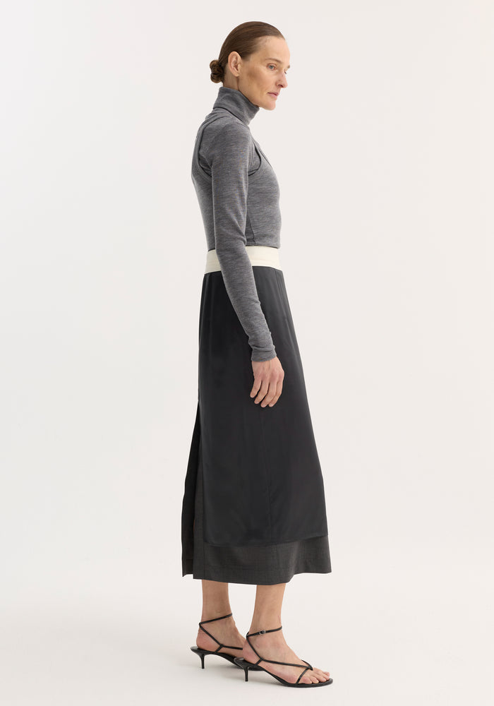 Model in profile wearing a mid grey melange double-layer wool top, featuring a fitted turtleneck and tank combination, paired with a layered skirt and sandals.