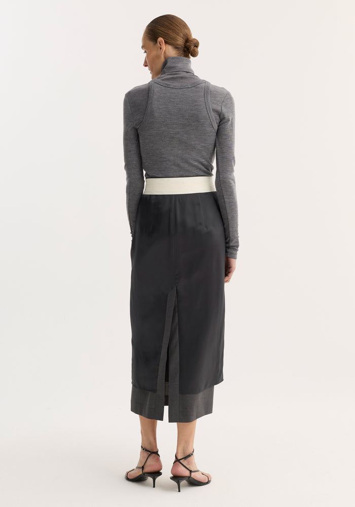 Woman wearing Double-layer wool top in mid grey melange, cinched with an off-white belt, over black satin skirt and grey trousers, styled with low bun.