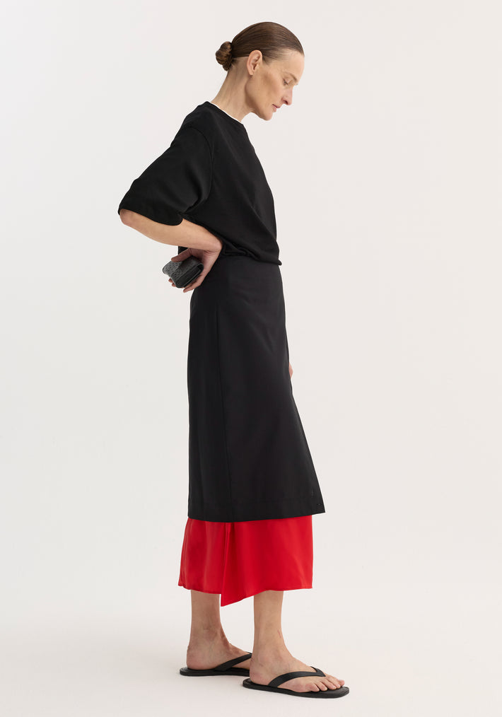 Model in profile wearing a black Contrast upside-down skirt with a red underlayer, holding a black clutch behind back, styled with a minimalist top and sandals.