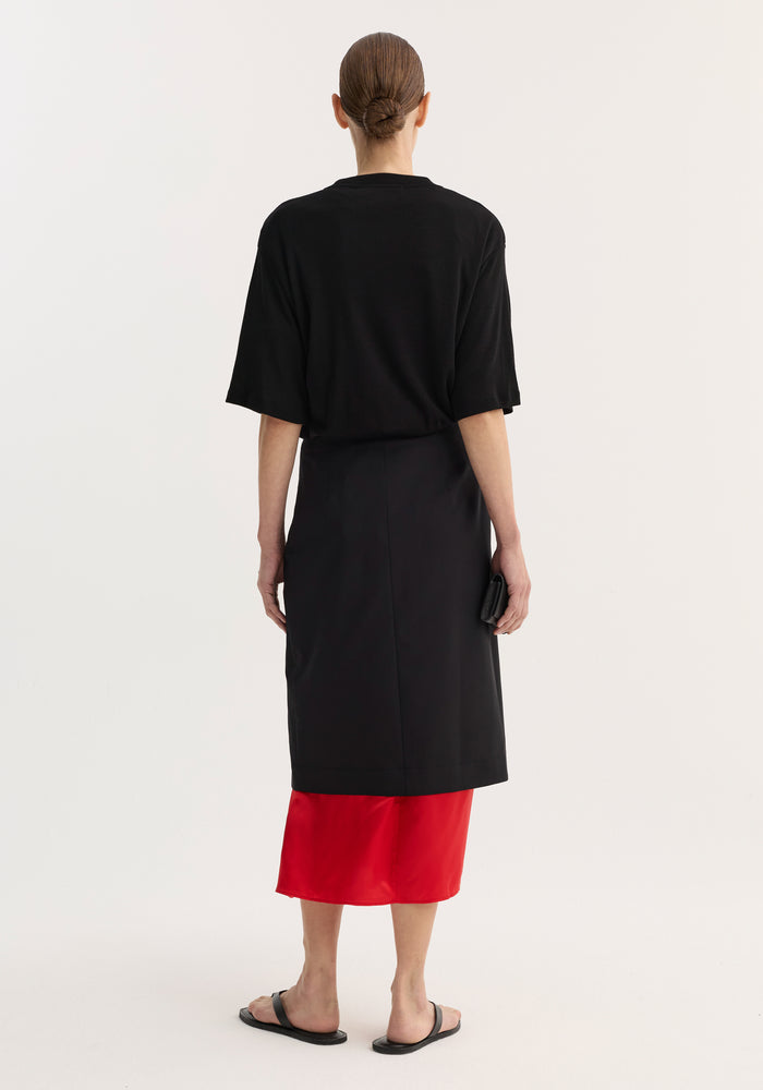 Contrast upside-down skirt | black, shown from the back, featuring a tapered silhouette with a front slit and a bold red underlayer, worn with black sandals.