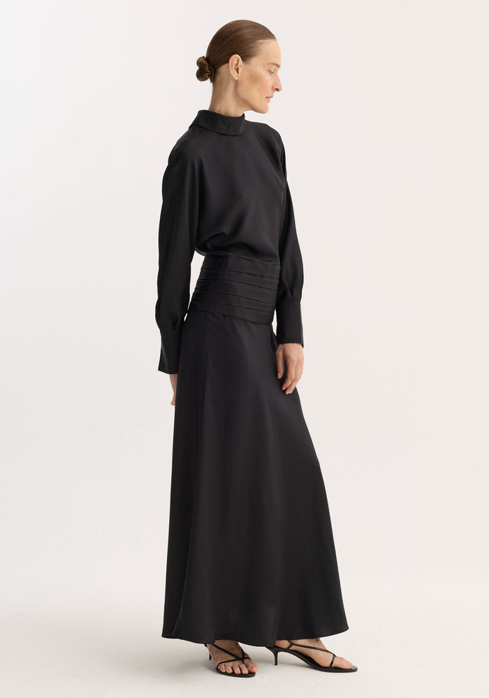 Model wearing Tuxedo silk dress | black, featuring long sleeves, mock-neck, pleated waist, and strappy sandals. Hair in bun, profile pose, hands at sides.
