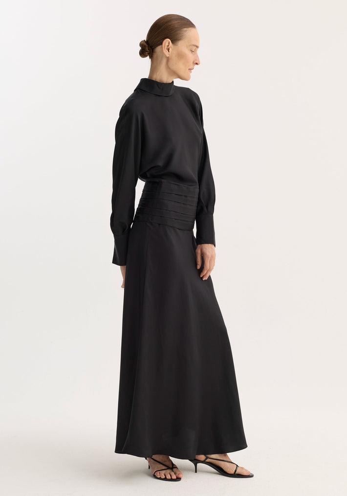 Model wearing Tuxedo silk dress | black, featuring long sleeves, mock-neck, pleated waist, and strappy sandals. Hair in bun, profile pose, hands at sides.