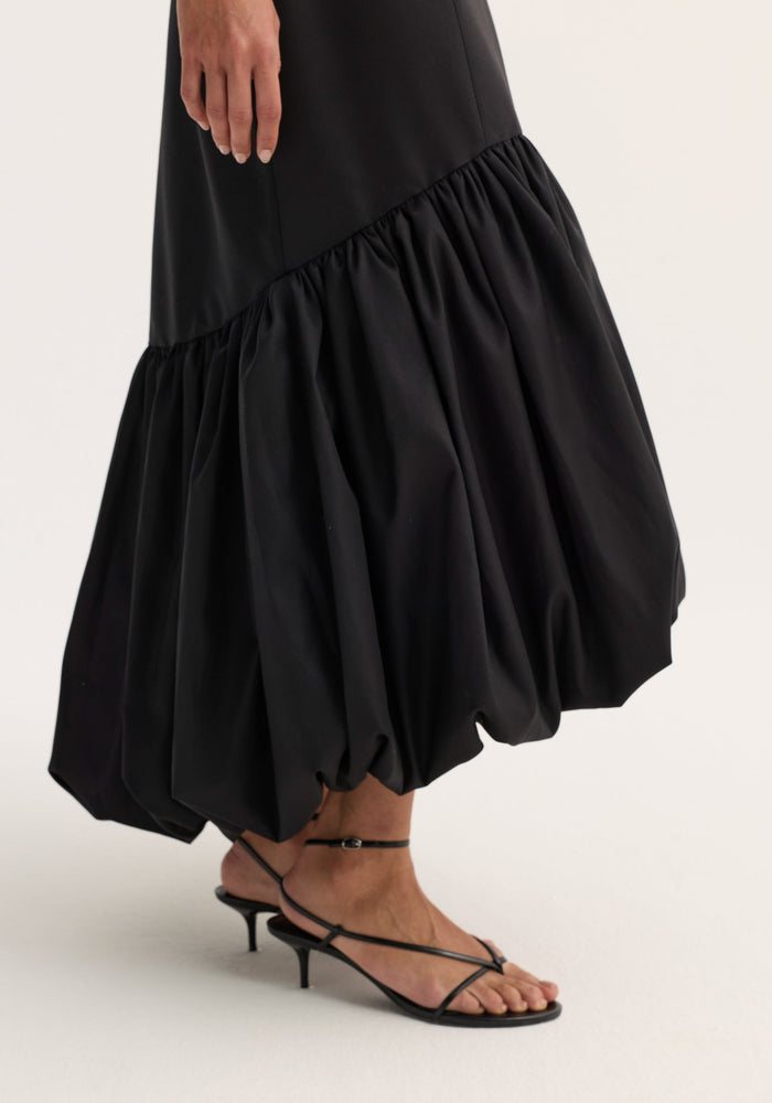 A person wears a Voluminous dress | black with a balloon-style hem and strappy sandals, showcasing the dress's sculptural movement and refined fit.