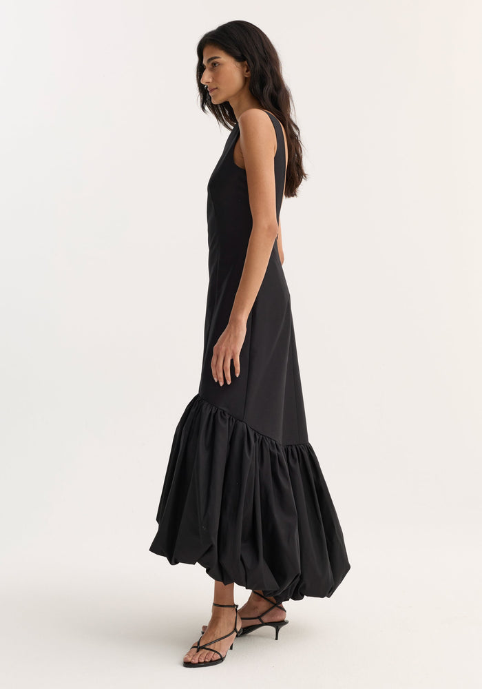 Model in Voluminous dress | black, showcasing a fitted, sleeveless wool-silk design with a boat neckline and a sculptural peplum hem, paired with strappy sandals.