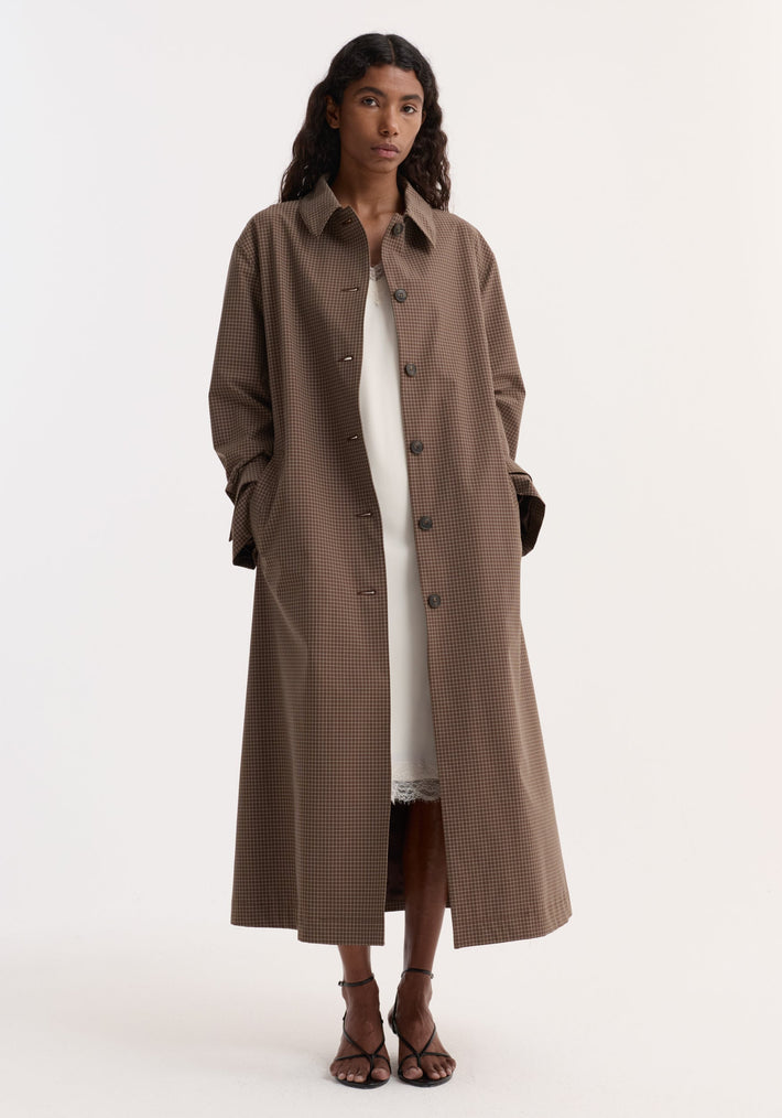 Model in Technical check belted coat, brown multi check, with a tie belt, structured collar, and cuff straps, over a white slip dress.