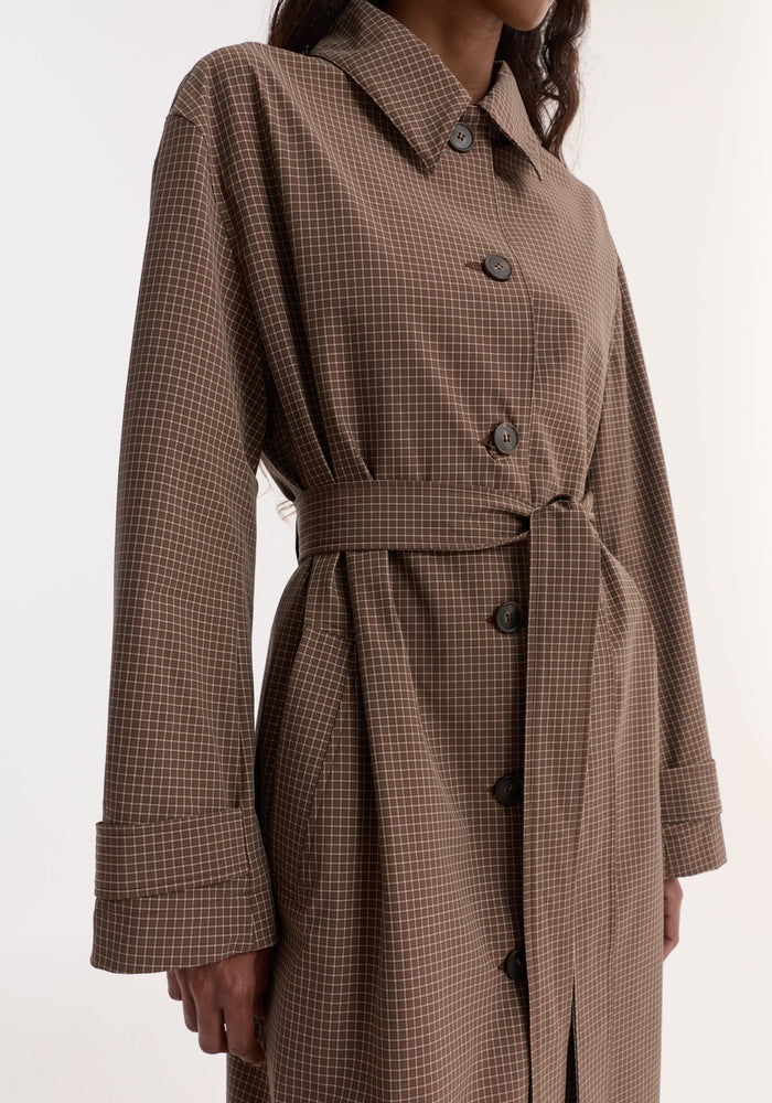 Technical check belted coat in a brown multi-check pattern, featuring a pointed collar, large buttons, cuff straps, side pockets, and a waist-defining tie belt.