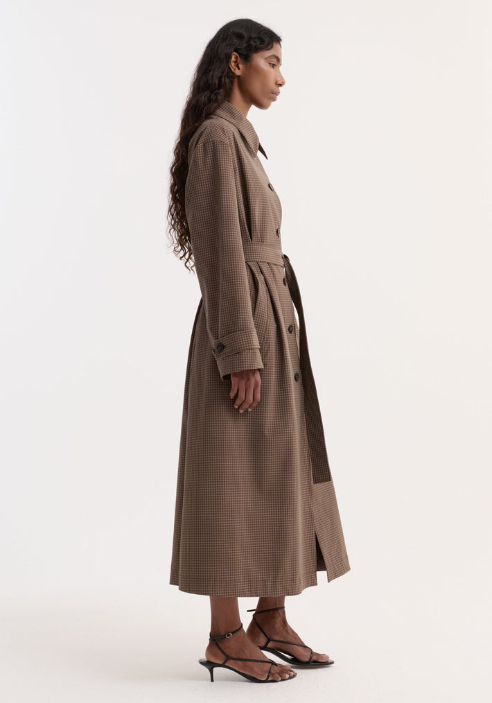 Technical check belted coat in brown multi check, featuring a structured collar, tie belt, cuff straps, and slanted pockets, worn with black sandals.