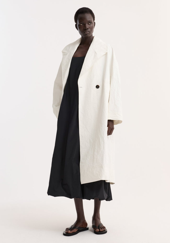 Model wears an oversized crushed cotton coat with raglan sleeves, wide lapels, and a single black button, over a black strapless dress and flat sandals.