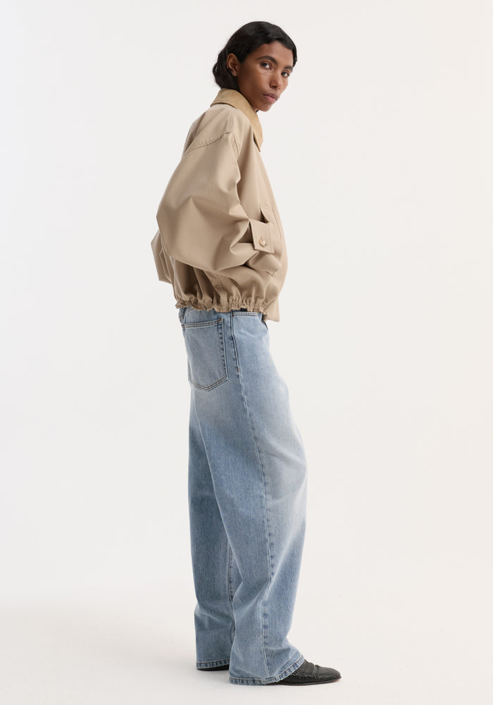 Person in profile wearing a Suede collar blouson jacket | sand, showcasing cropped trench-inspired design with wide sleeves and suede collar, hands in pockets.