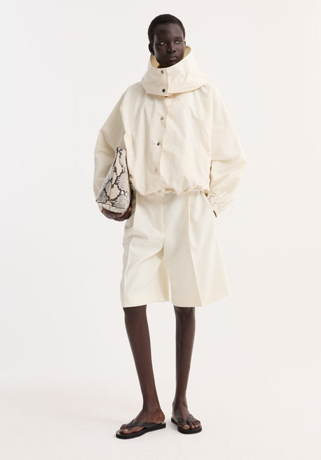 Volume silk-blend jacket in cream, oversized with hood and snap closures, worn by model holding a snakeskin tote, paired with matching shorts and black sandals.