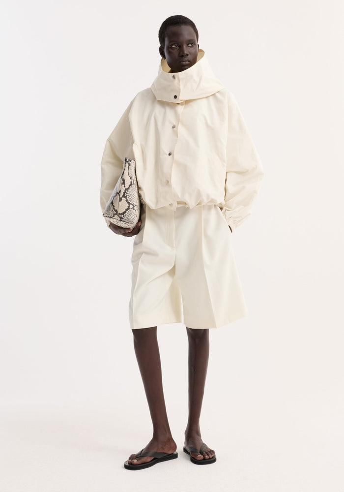 Volume silk-blend jacket in cream, oversized with hood and snap closures, worn by model holding a snakeskin tote, paired with matching shorts and black sandals.