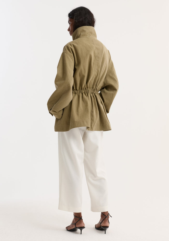 Model in Sculptural safari jacket | sage, with a stand collar, oversized bellows pockets, and elasticated waist, paired with wide-leg trousers and low-heel sandals.