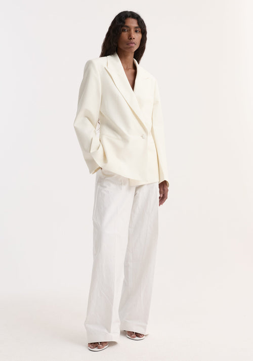 Model in Hourglass shaped blazer, off white, featuring a sharp notch lapel, cinched waist, single button, paired with wide-leg trousers and open-toe sandals.