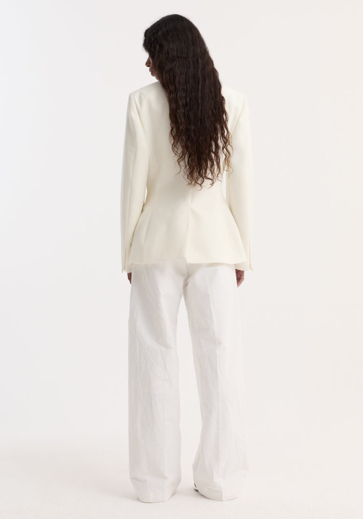 Person wears Hourglass shaped blazer | off white featuring sharp notch lapel, cinched waist, peplum hem, and single button, paired with loose white trousers.