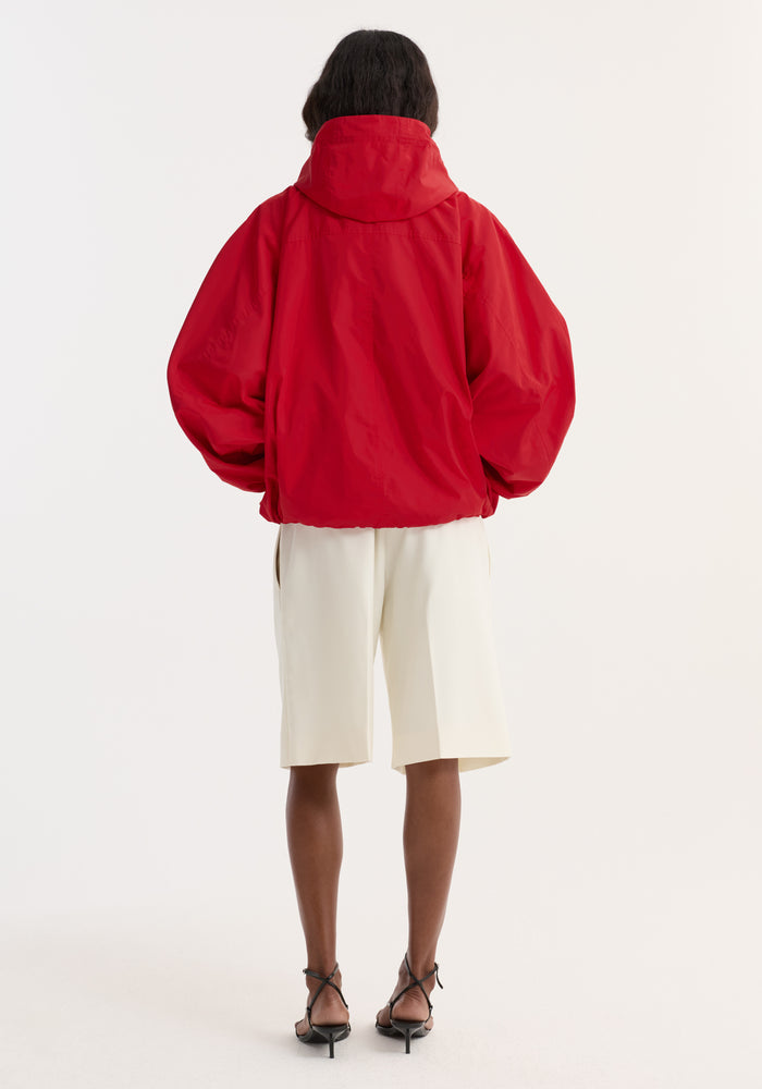 Volume silk-blend anorak | red features a hood, voluminous sleeves, front pocket, and zip side pockets. Styled with cream shorts and black sandals.