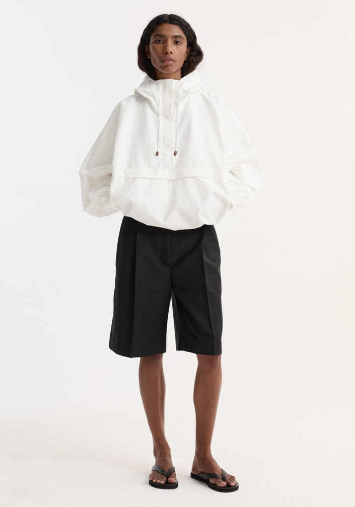 Model wearing an oversized optic white textured cotton-blend anorak with hood, front and side pockets, paired with black knee-length shorts and flip-flop sandals.